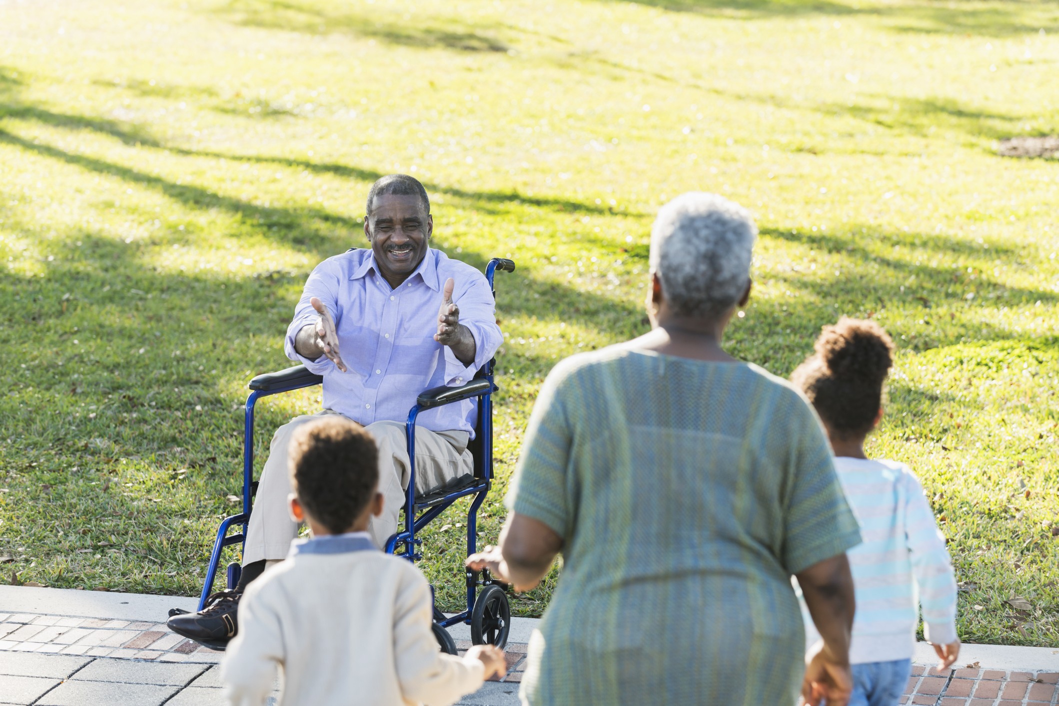 A Black grandfather in a wheel chair holding out his arms to greet his grandson and granddaughter for hugs along with his wife